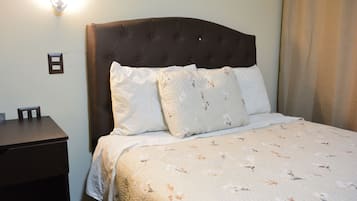 Una cama bien hecha con una colcha estampada con motivos florales, un cabecero tapizado oscuro y una mesita de noche con una lámpara.