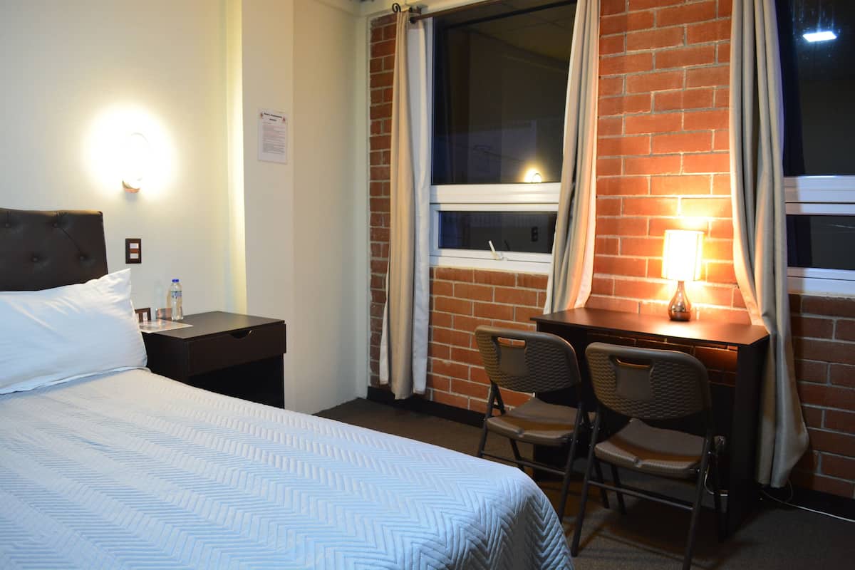 A hotel room with a bed, a desk, two chairs, a window with curtains, and a wall-mounted lamp.