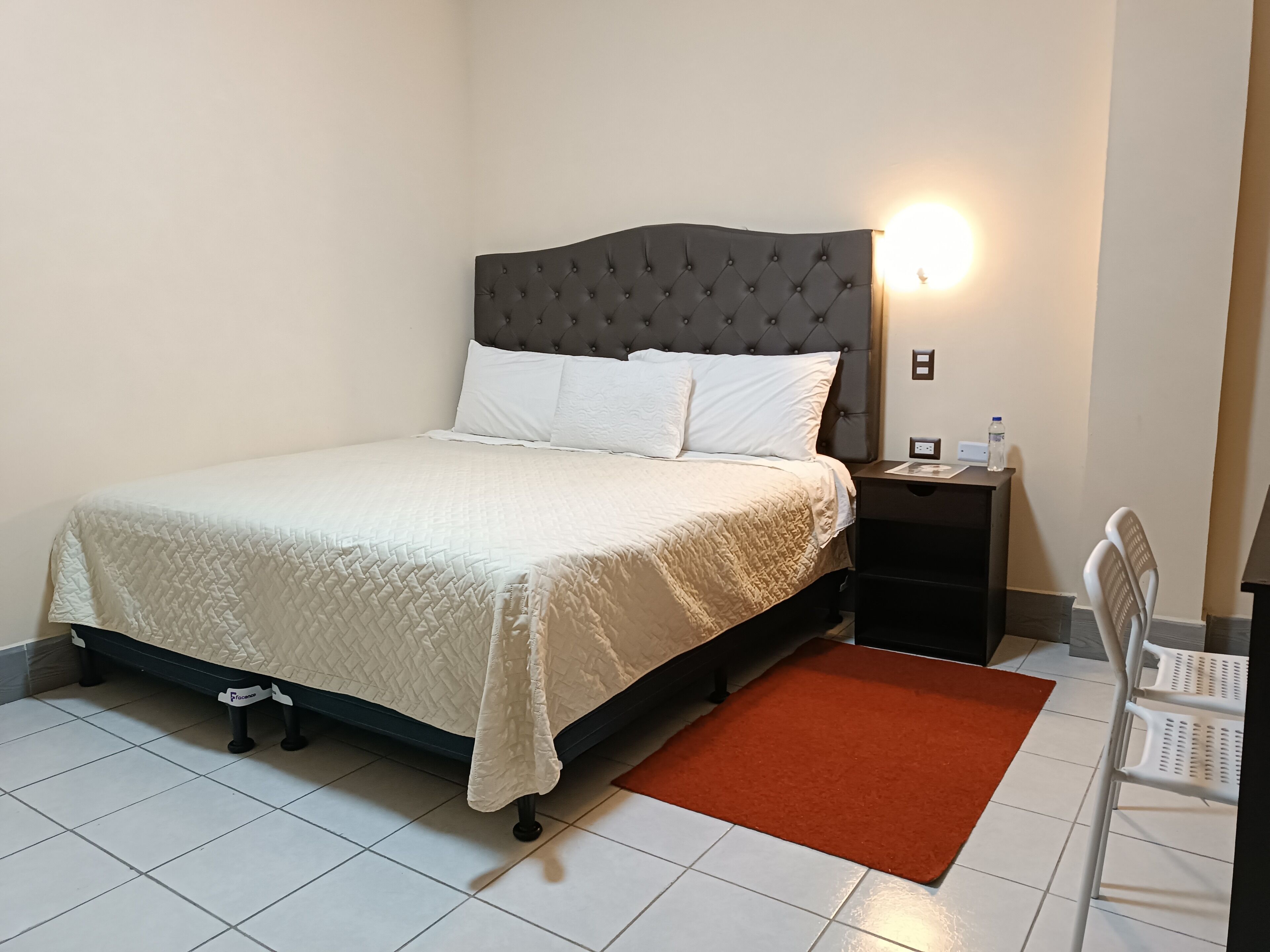 Una cama bien hecha con cubrecama acolchado, una mesita de noche con lámpara y una alfombra roja sobre suelo de baldosas.