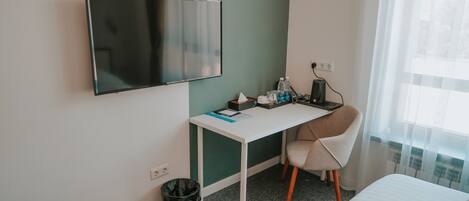 A hotel room with a white desk, a chair, a bed, a TV mounted on the wall, a trash bin, and a window with curtains.