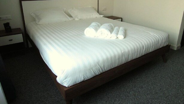 A neatly made bed with white linens and rolled towels, a wooden headboard, a bedside table with a lamp, and a plain wall. - Hotel 78 Cafe (Karakol)