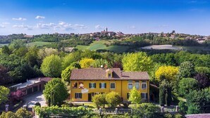 Una villa amarilla con techo de tejas, rodeada de abundante vegetación y vista a un pueblo lejano con una torre destacada.