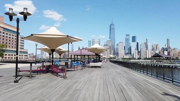 Une promenade en bois agrémentée de bancs et de parasols, avec la silhouette de la ville en arrière-plan et une rivière.