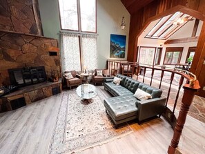 A spacious living room with a stone fireplace, a large sectional sofa, a coffee table, and a view of the outdoors through large windows.