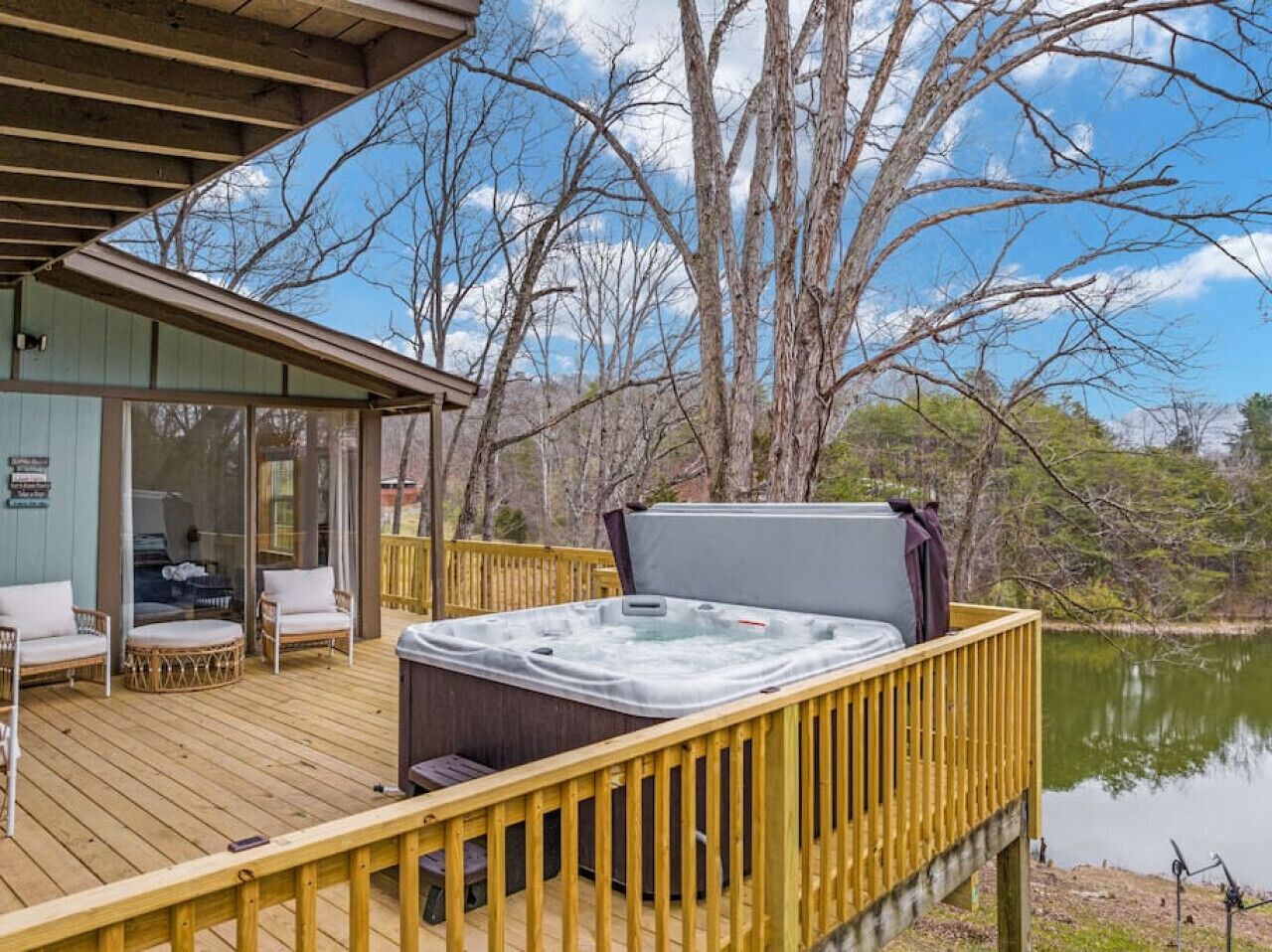 A wooden deck with a hot tub, outdoor seating, and a view of a lake surrounded by trees.