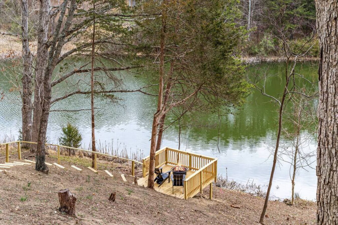 A wooden deck with a fire pit and a bench overlooking a calm lake surrounded by trees.