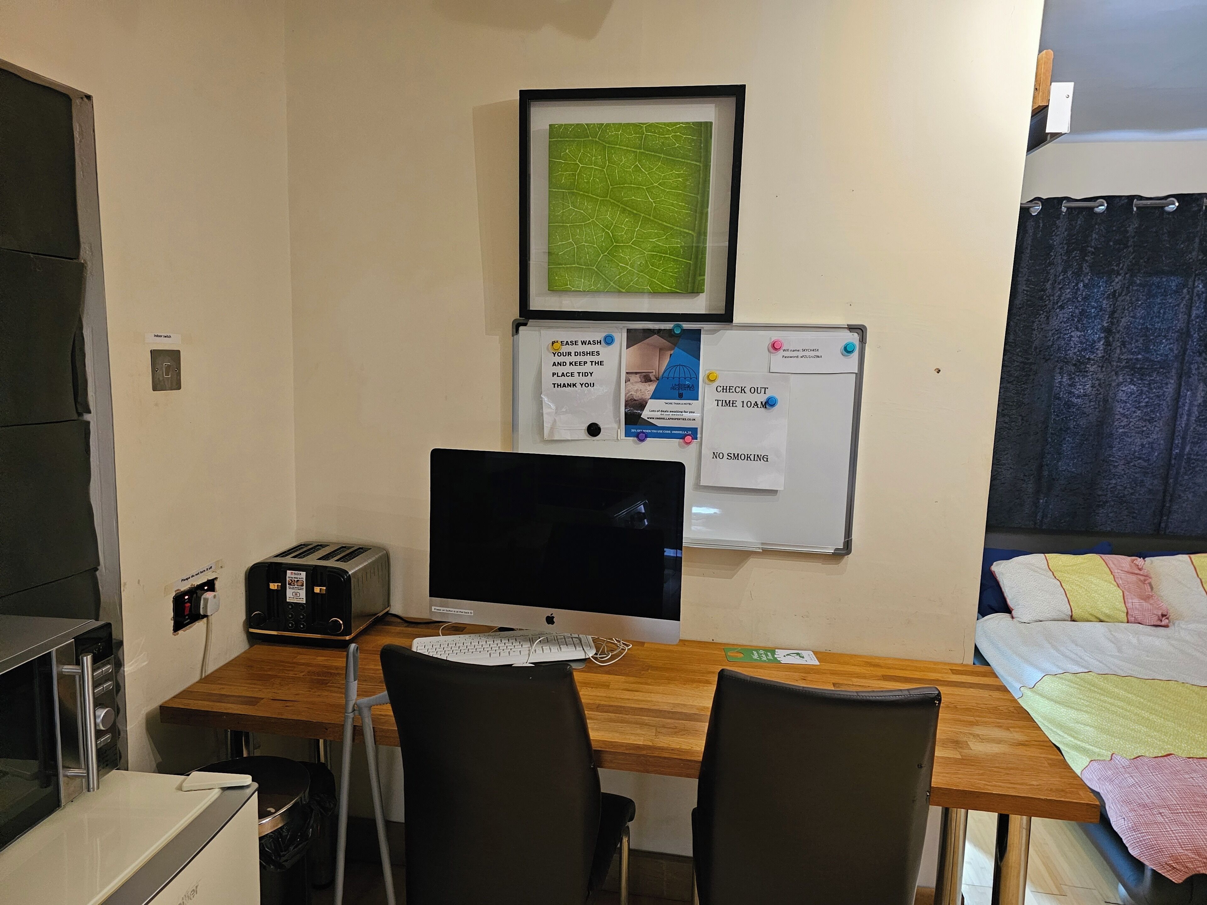 A small, tidy room with a wooden desk, a computer, a microwave, and a bed with a colorful blanket.
