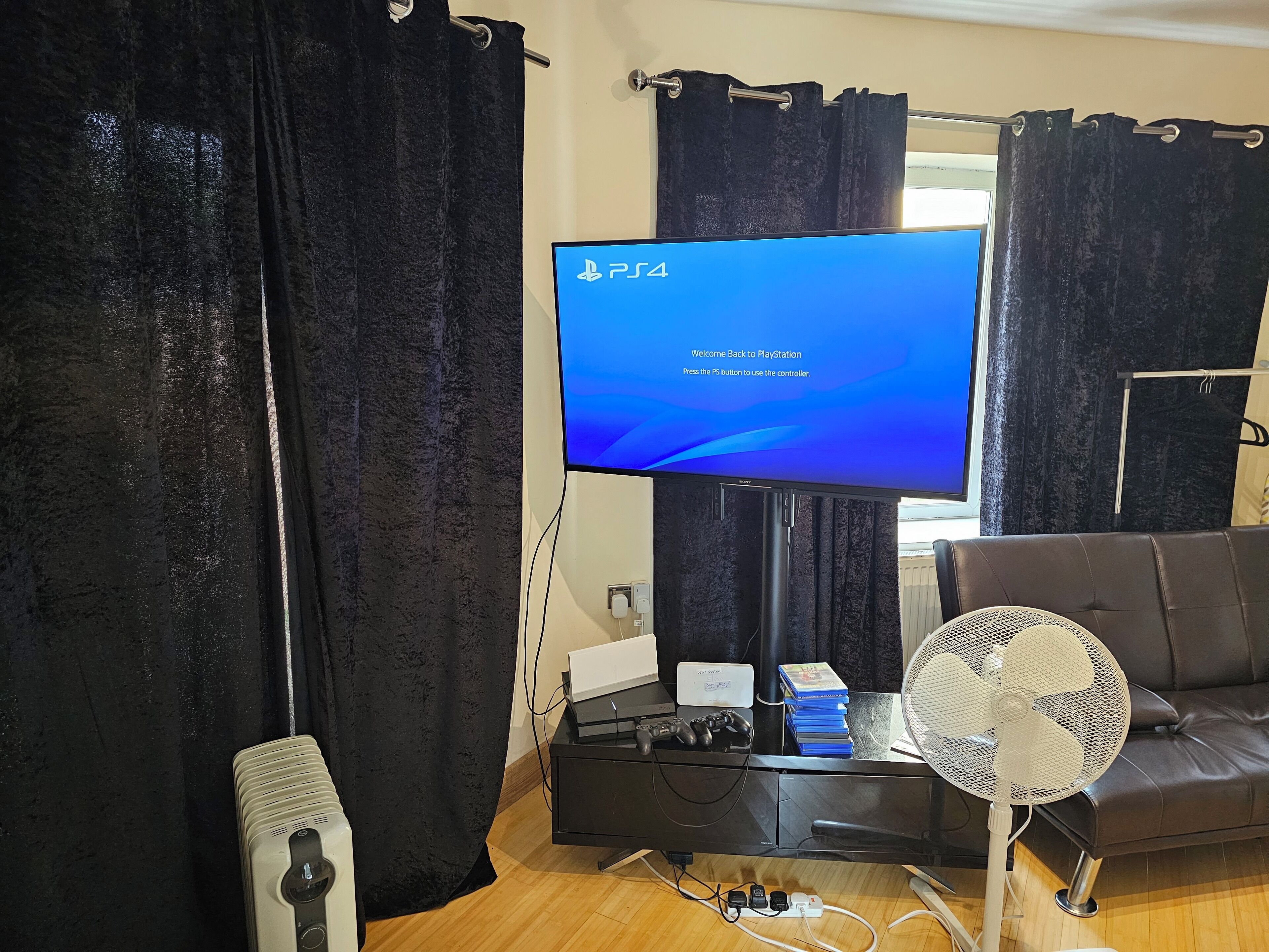 Une console PlayStation 4 sur un écran de télévision, accompagnée d’un message, un meuble noir contenant des jeux et un ventilateur, et un canapé en cuir.