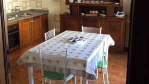 Un comedor con una mesa lista para cuatro personas, rodeada de sillas blancas con cojines verdes. En el fondo, un aparador de madera con vajilla y un reloj en la pared.