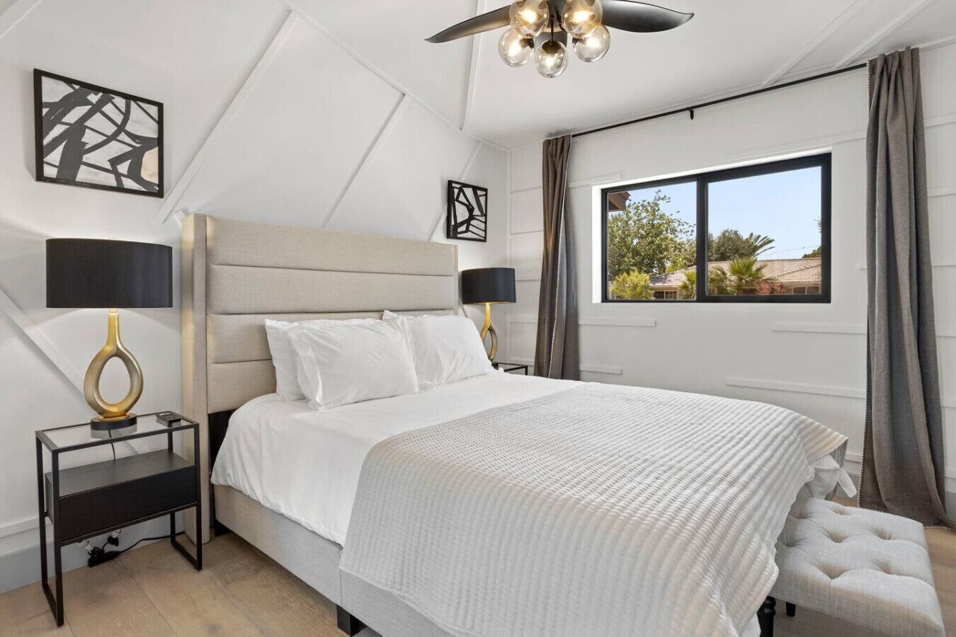 A modern bedroom with a large bed, two bedside tables with lamps, a ceiling fan, and a window with a view of greenery.