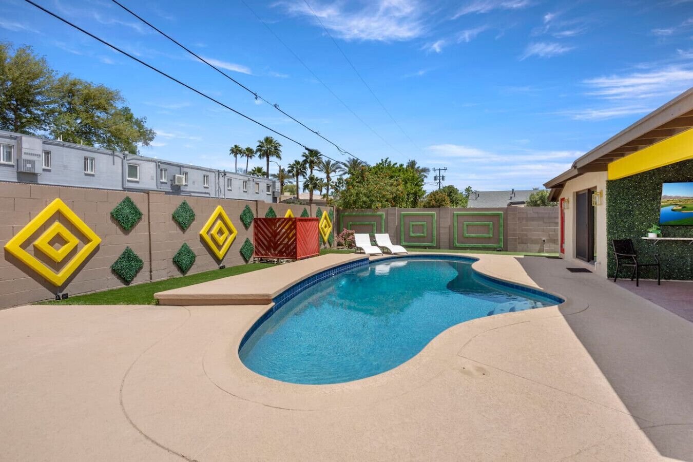 Una piscina de diseño curvo, rodeada por una terraza color beige. Hay sillas de descanso y una pequeña mesa con sillas a la derecha. La zona de la piscina está delimitada por un muro con diseños decorativos y palmeras al fondo.