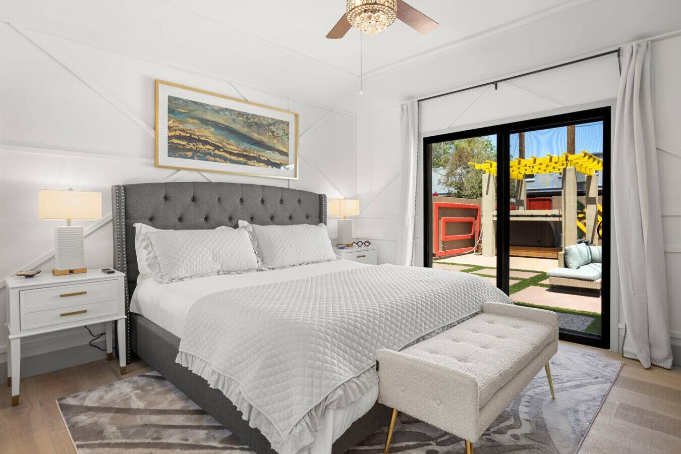 A modern bedroom with a large bed, a nightstand, a bench, and a view of the outdoors through a sliding glass door.