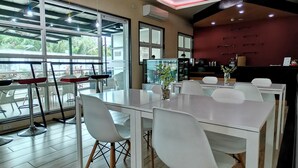 A modern dining area with a long table, white chairs, and a view of the outdoors through large windows. - La Piazza Hotel and Convention Center  (Legazpi)