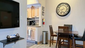 Un espacio habitable compacto con kitchenette, una zona de comedor con mesa y sillas, un reloj montado en la pared y un televisor.