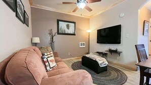 Una sala de estar con un sofá rosa, un otoman negro, un ventilador de techo, un televisor de pantalla plana y un cuadro en la pared.