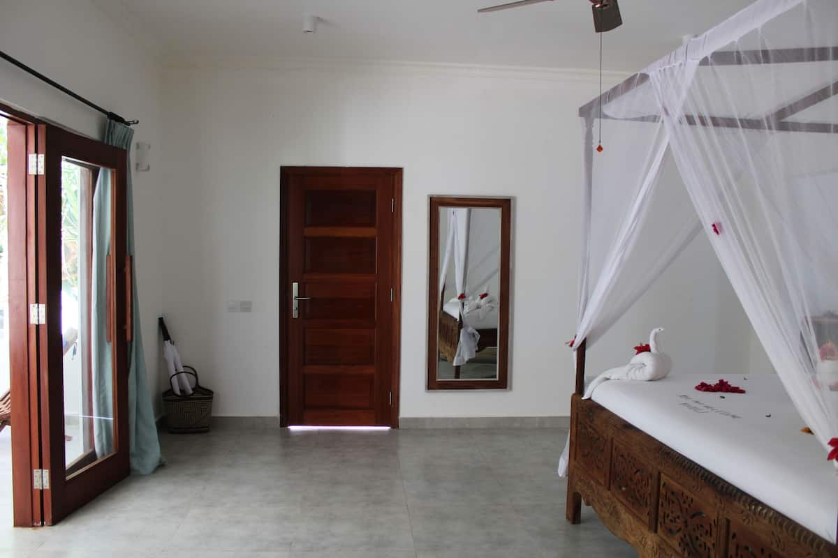 Un dormitorio con una cama de madera, un ventilador de techo, una puerta de madera, un espejo y una ventana con cortinas.