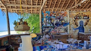 A beachside bar with a rustic wooden ceiling, a mural, and a variety of bottles and glasses. - Kivuli Beach Resort Paje (Paje)