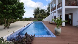 A rectangular swimming pool with a tiled edge, surrounded by a wooden deck and a white building with balconies. - Kivuli Beach Resort Paje (Paje)
