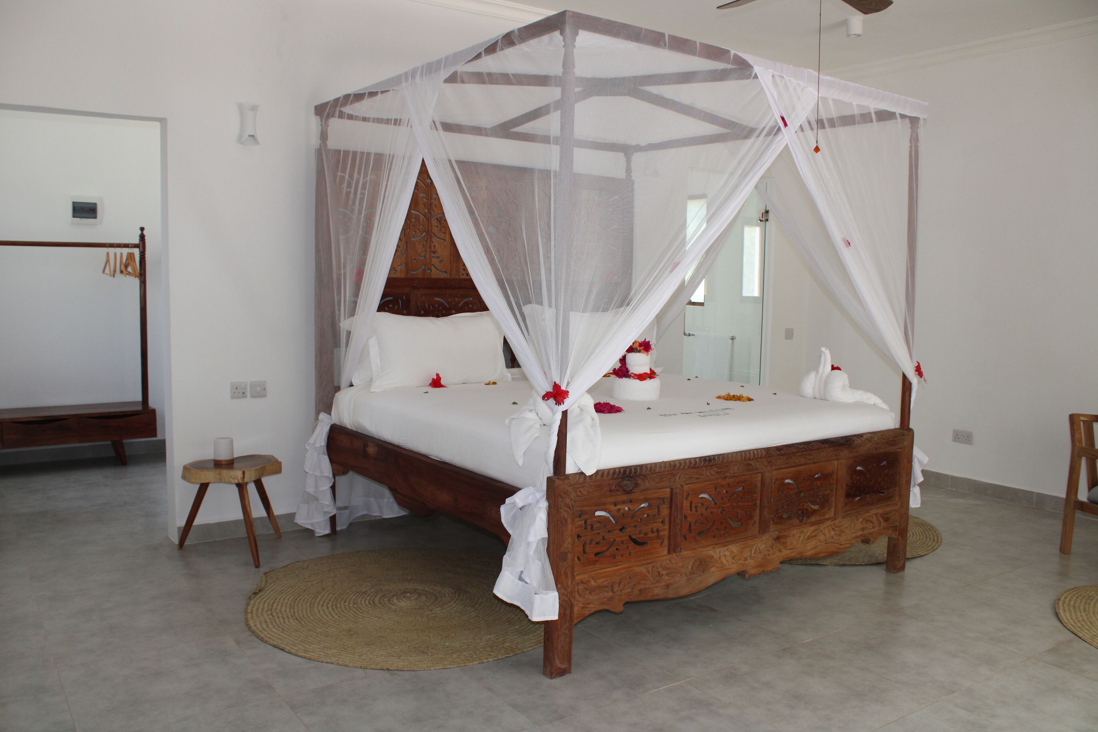 A four-poster canopy bed with a wooden frame and white bedding, a small round wooden stool, and a woven mat on the floor.