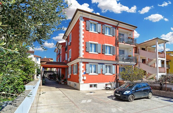 A multi-story building with balconies, a car parked in front, and a clear sky.