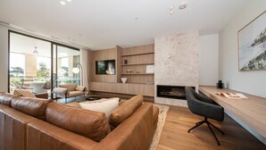 Een moderne woonkamer met een leren bank, een houten salontafel en een zwarte bureaustoel. Er is een open haard, een aan de muur bevestigde televisie en een balkon met tuinmeubilair.