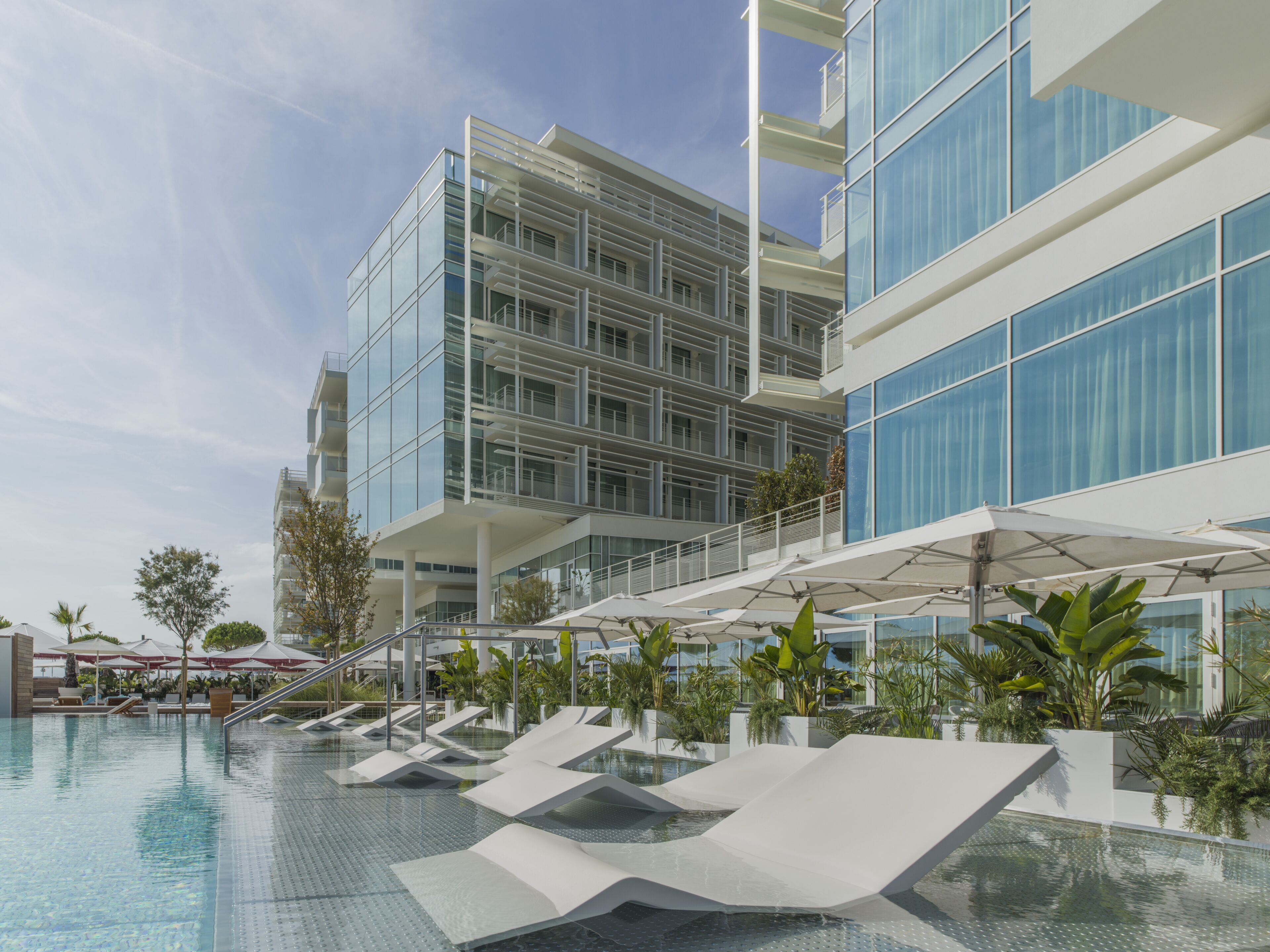 Un hotel moderno con fachada de vidrio, zona de piscina con sillones de descanso y cielo despejado.