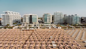 Un hôtel situé en bord de mer, composé de plusieurs immeubles de grande hauteur, doté d’une vaste plage de sable avec de nombreux parasols et chaises longues, ainsi que d’un espace piscine.