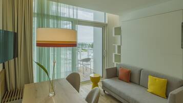 A modern living room with a wooden dining table, a sofa with cushions, a floor lamp, and a view through a large window to the outdoors.