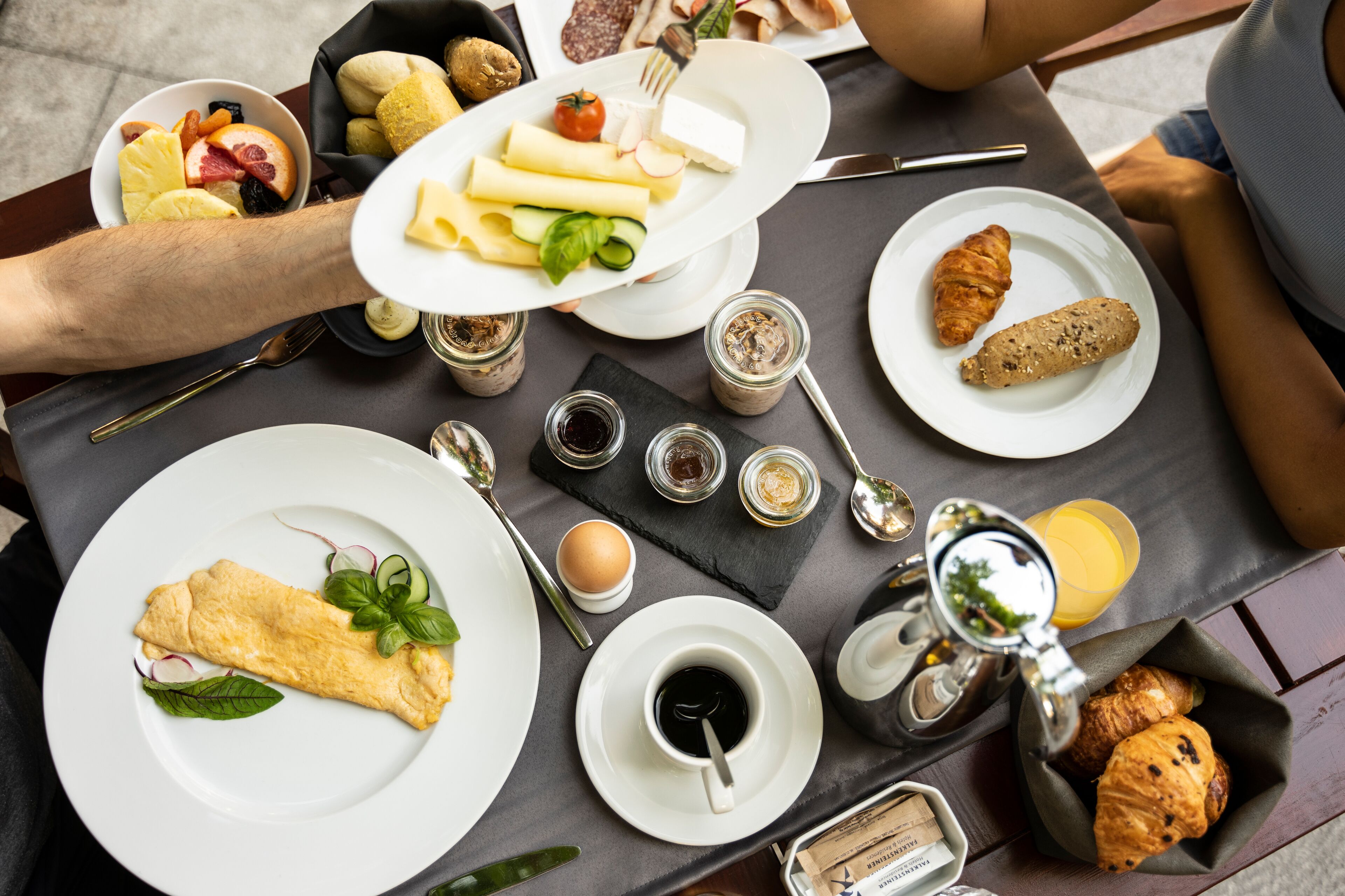 Une table dressée avec des aliments pour le petit-déjeuner, notamment des œufs, des croissants et une omelette roulée, ainsi que des condiments et une machine à café.
