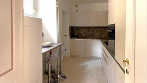 A narrow kitchen with white cabinets, a marble backsplash, and a small dining area with a glass table and chair. - Cà del Conte Lake Como (Tremezzo)