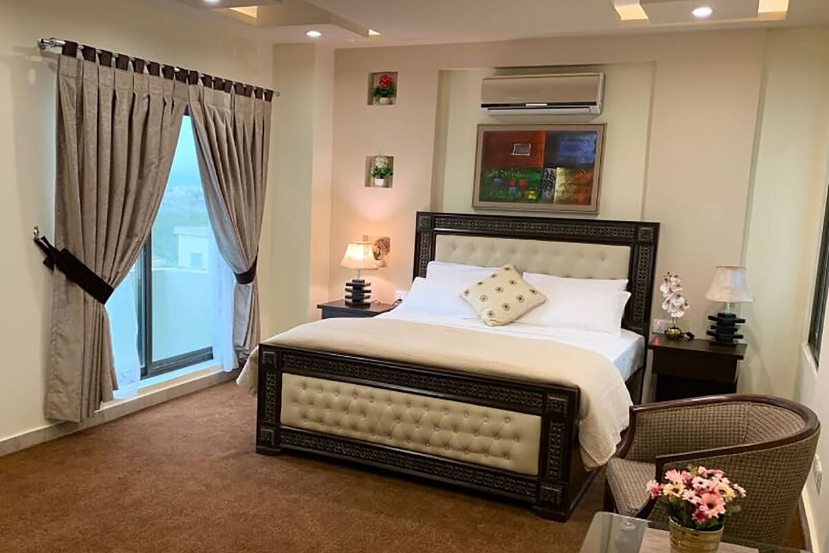 Ein Schlafzimmer mit einem großen Bett, einem Fenster mit Vorhängen, einer Wand-Klimaanlage und einem kleinen Tisch mit einer Lampe und Blumen.