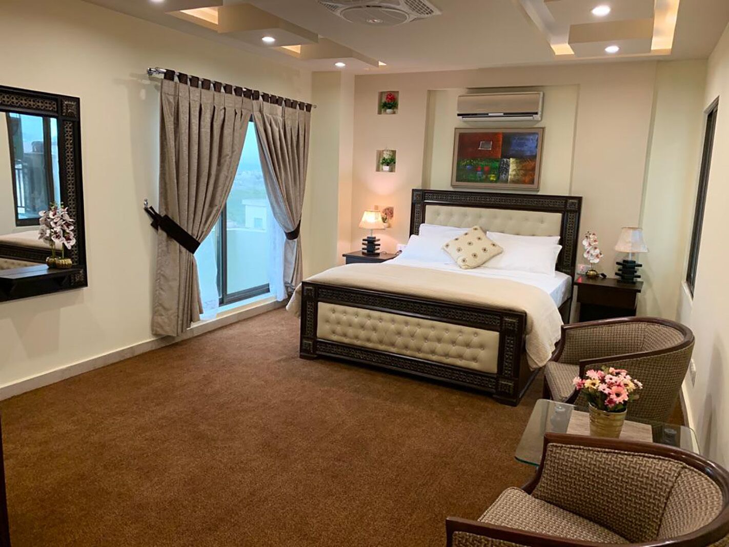 A bedroom with a large bed, a window with curtains, a nightstand with a lamp, a wall-mounted air conditioner, and a mirror.