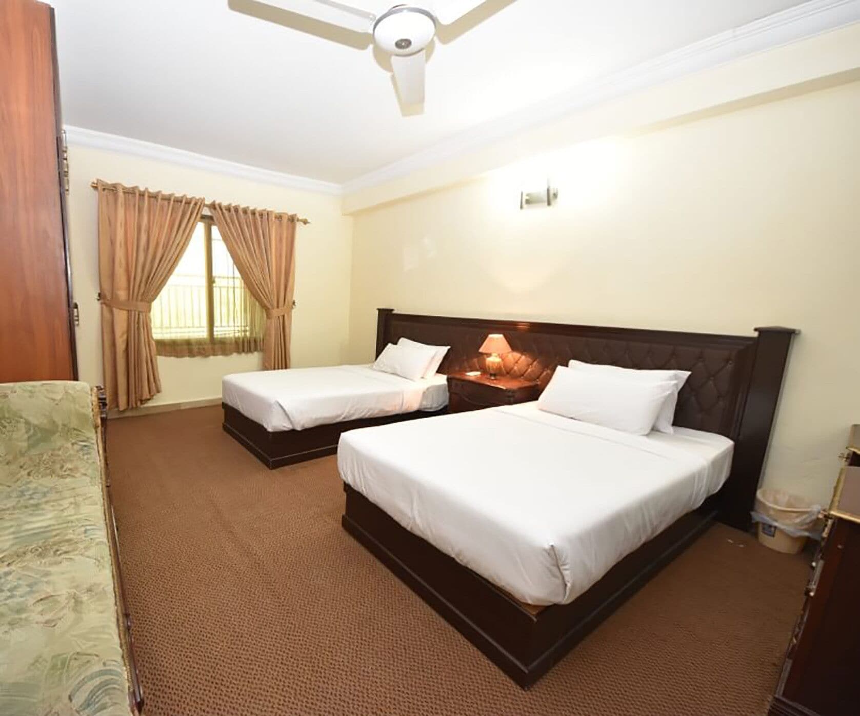Habitación de hotel con dos camas, ventilador de techo, ventana con cortinas y mesita de noche con lámpara.
