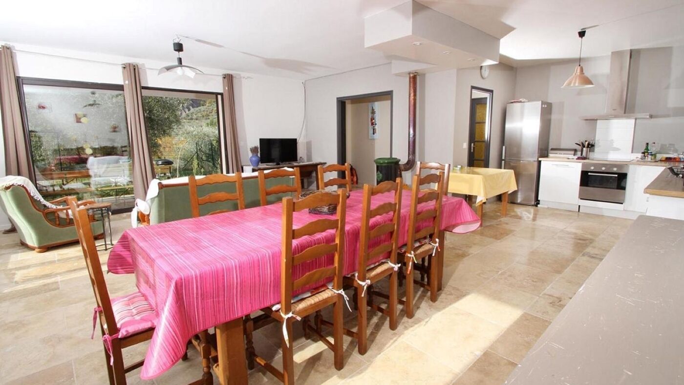 Comedor con mesa dispuesta para seis personas, sillas de madera, televisor y zona de cocina con refrigerador y horno.