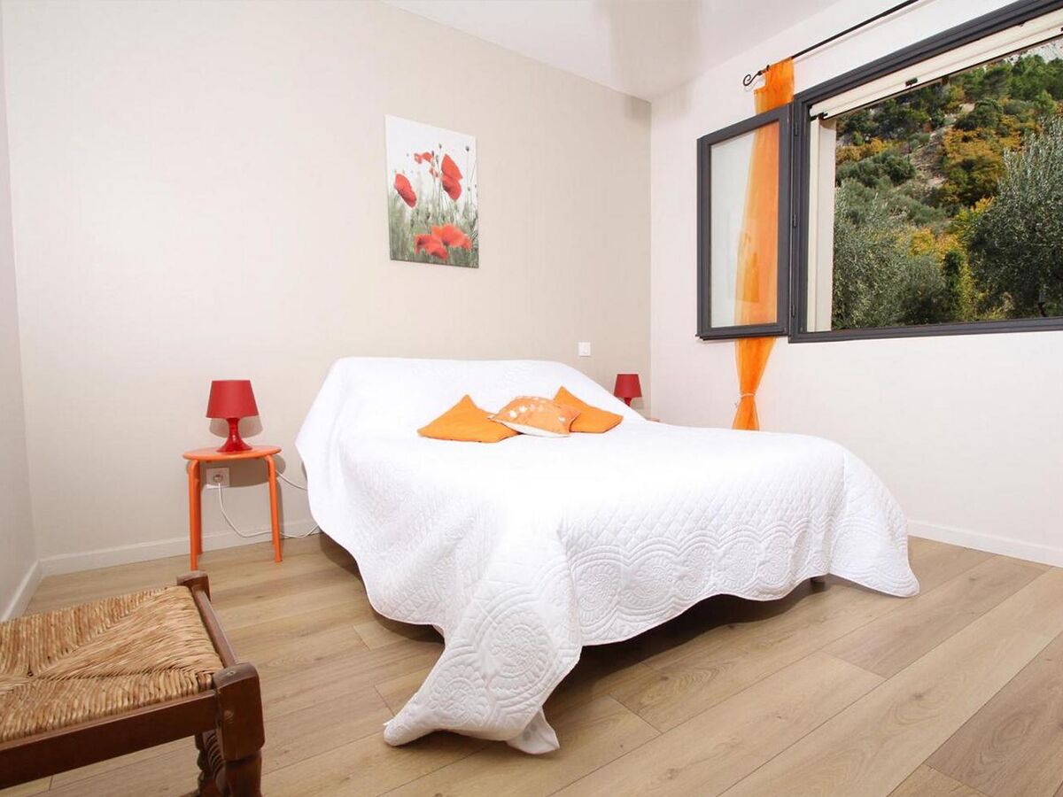 Un dormitorio con una cama, una mesita de noche con una lámpara, una silla y un ventanal con vistas al verdor.