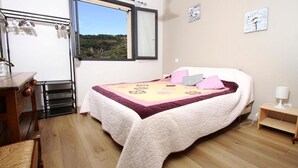 A bedroom with a bed, a wooden dresser, a desk, a chair, a window with a view of the outdoors, and a small table with a lamp. - Villa des Oliviers (Buis-les-Baronnies)