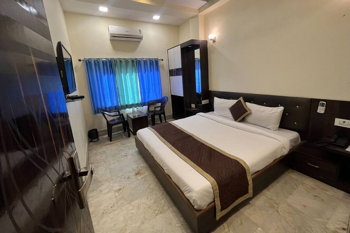 Quarto de hotel com uma cama grande, uma escrivaninha, duas cadeiras, aparelho de ar condicionado e uma televisão.