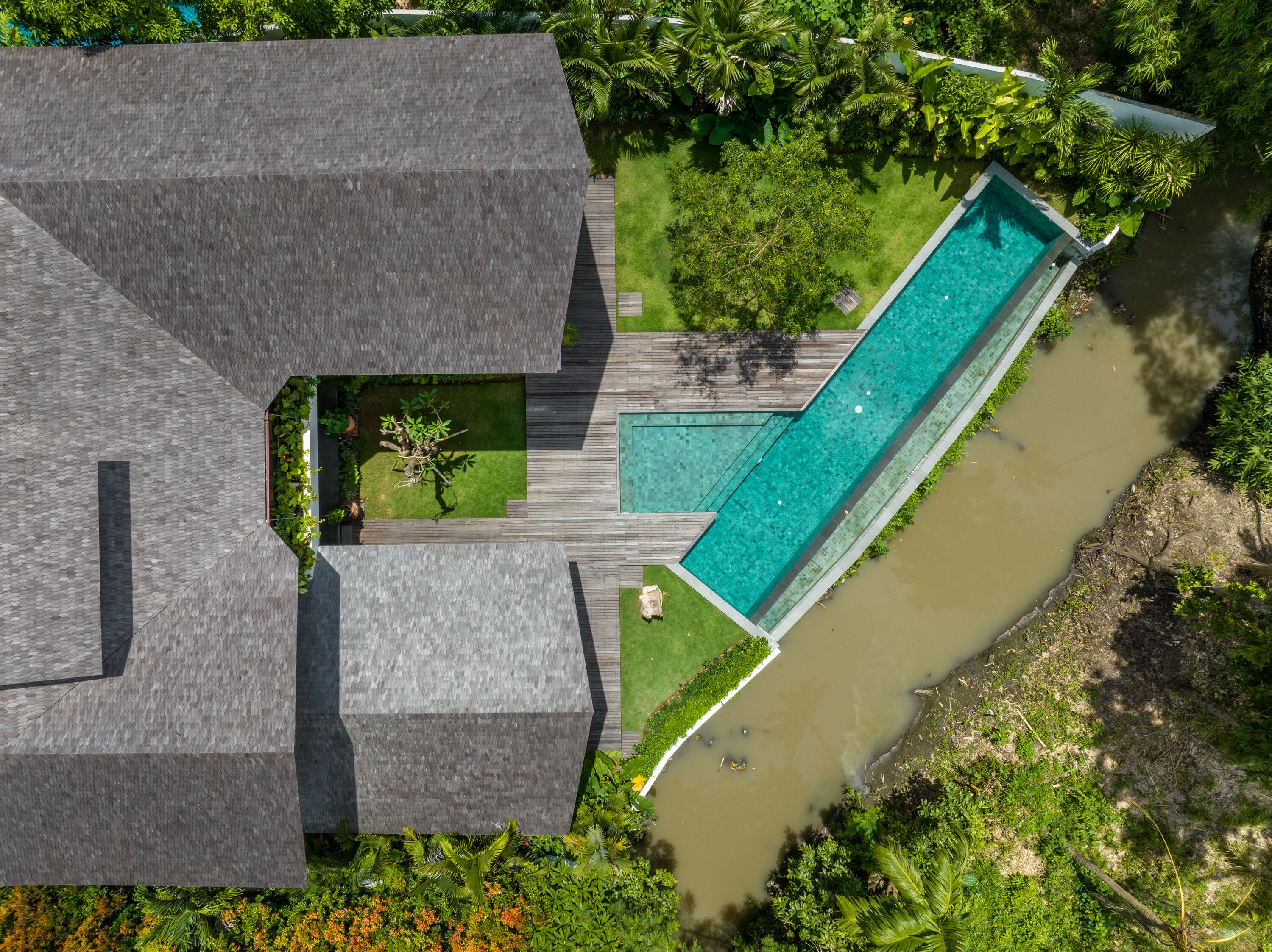 Luftaufnahme eines modernen Resorts mit einem langen Schwimmbecken, umgeben von üppiger Vegetation und einem Fluss.
