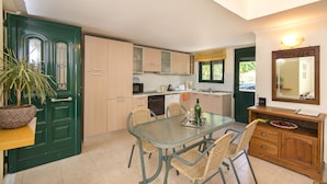 A compact kitchen with a dining area, featuring a green door, wooden cabinets, a tiled backsplash, and a dining table with chairs. - THERETRA KIOTARI VILLAS (Rhodes)