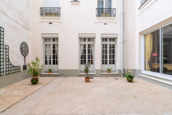 Une cour avec trois plantes en pot, un bâtiment blanc doté d’un balcon et une fenêtre avec un rideau.
