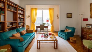 A living room with a teal sofa, a wooden bookshelf, a glass coffee table, and a view through a door with yellow curtains. - Postcards from the Eiffel Tower, by Veeve (Paris)