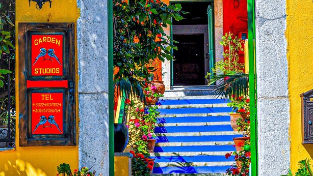A colorful entrance with a sign for 'Garden Studios' and a telephone number, leading to a staircase with blue steps and potted plants. - Symi Garden Studios (Symi)