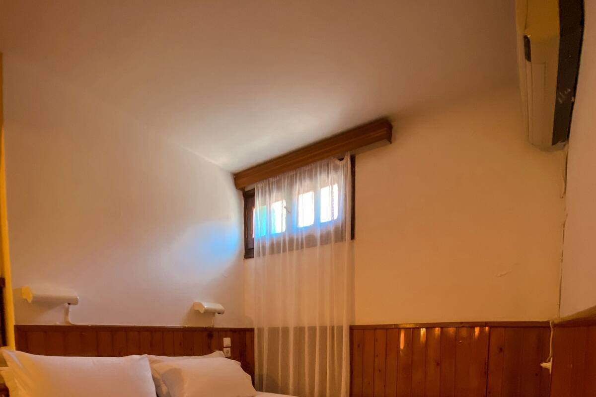 Ein ordentlich bezogenes Bett mit brauner Decke und weißen Kissen, ein hölzerner Kopfteil, ein Nachttisch mit Lampe und ein Fenster mit durchsichtigen Vorhängen.