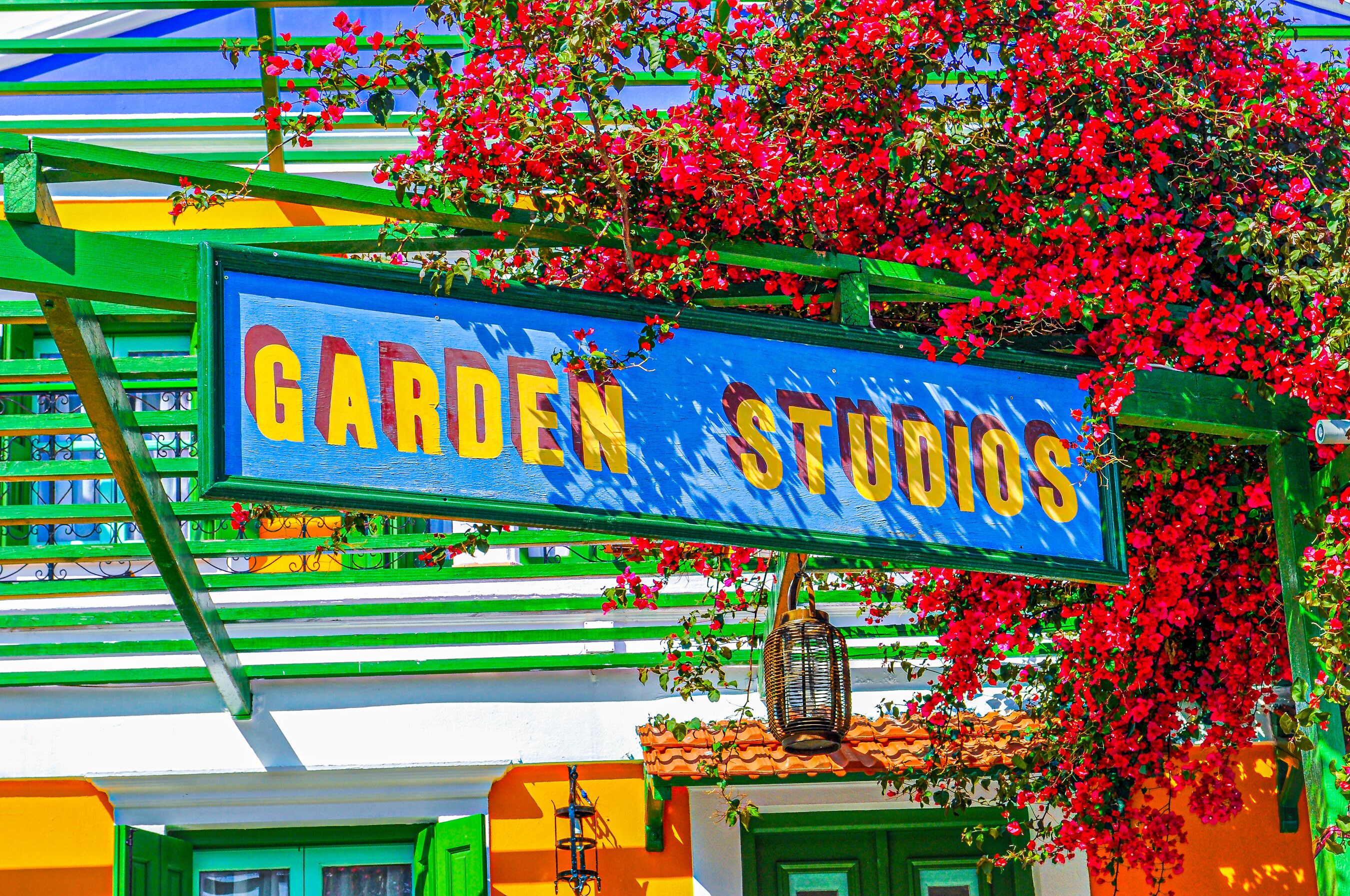 Un letrero colorido para 'Garden Studios' con una estructura verde y azul, flores rojas y un farol colgante.