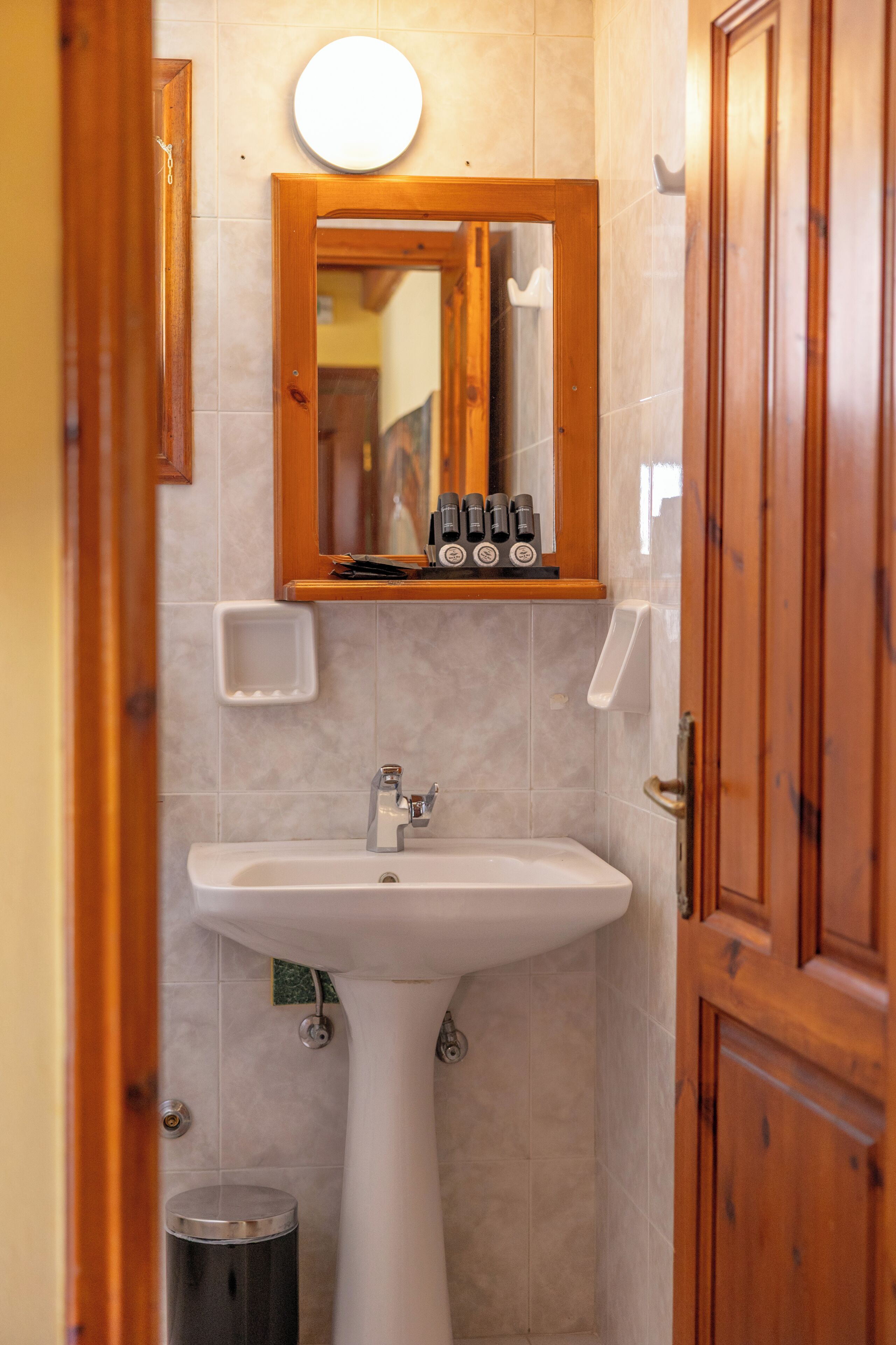 Un baño con un lavamanos pedestal, un espejo con marco de madera y un estante pequeño con artículos de aseo.