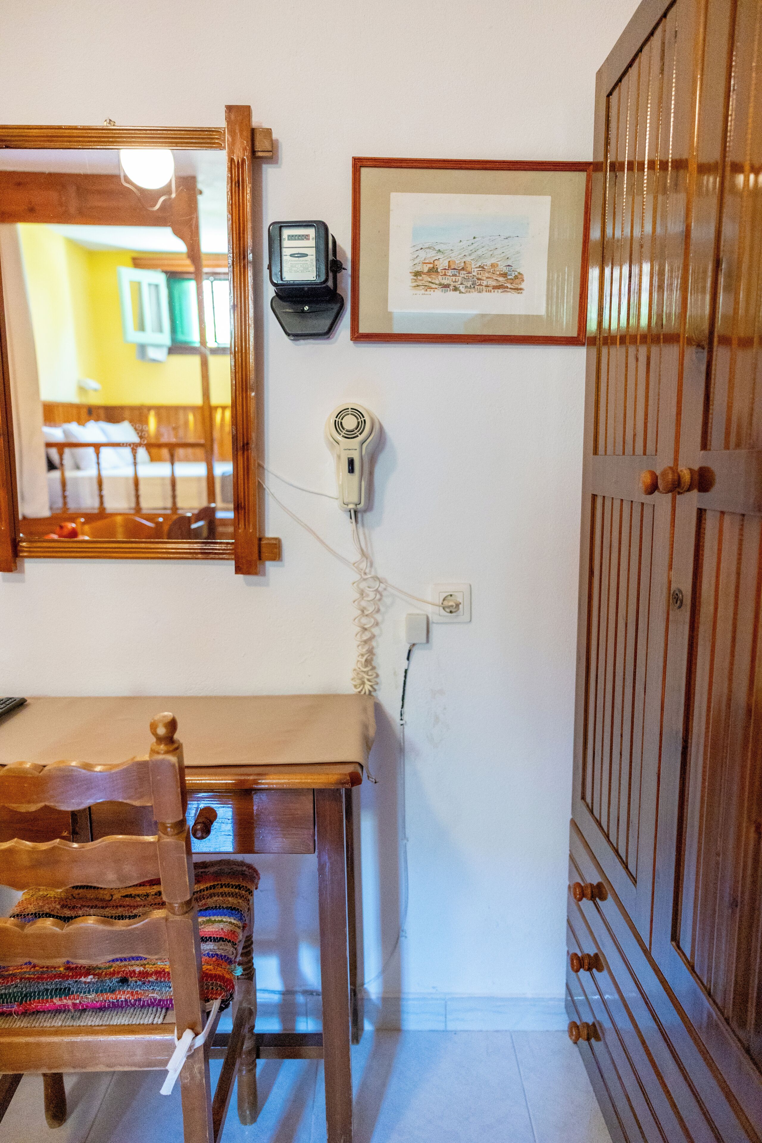 Una habitación con un escritorio de madera, una silla, un espejo, un secador de pelo, un cuadro enmarcado y un armario.