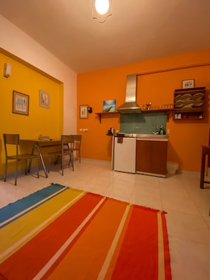 A compact kitchen with a stove, sink, and overhead ventilation. There are chairs and a dining table, and a colorful rug on the floor. - Symi Garden Studios (Symi)
