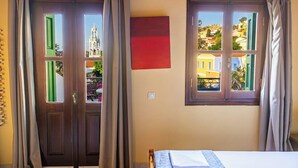 A room with two windows, a bed, a red wall piece, and a door. - Symi Garden Studios (Symi)