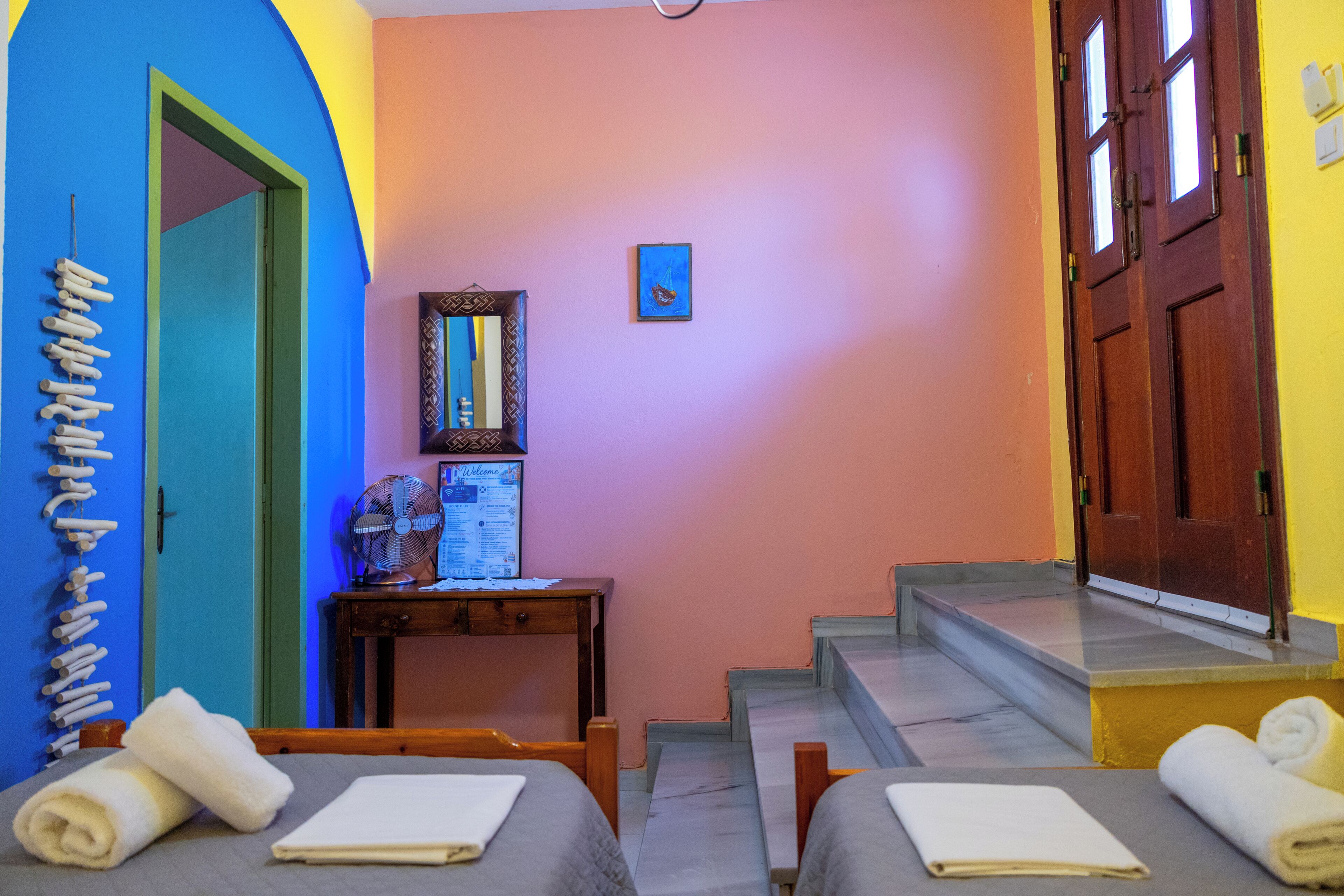 A room with two beds, a wooden desk, a small table with a fan, a framed picture on the wall, and a staircase leading to another room.