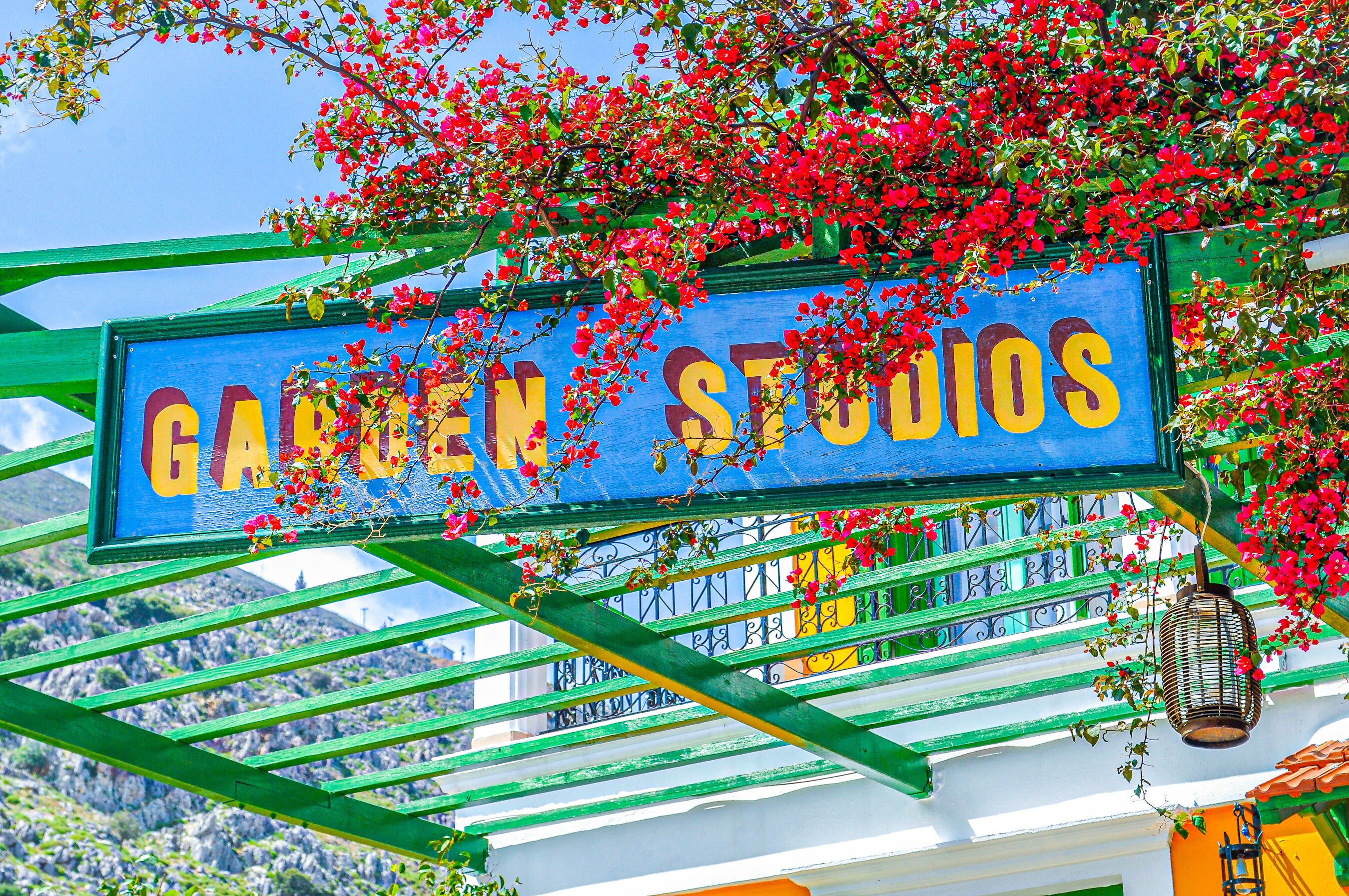 Et skilt for 'Garten Studios' med en grøn metalramme og blå baggrund, omgivet af røde blomster.